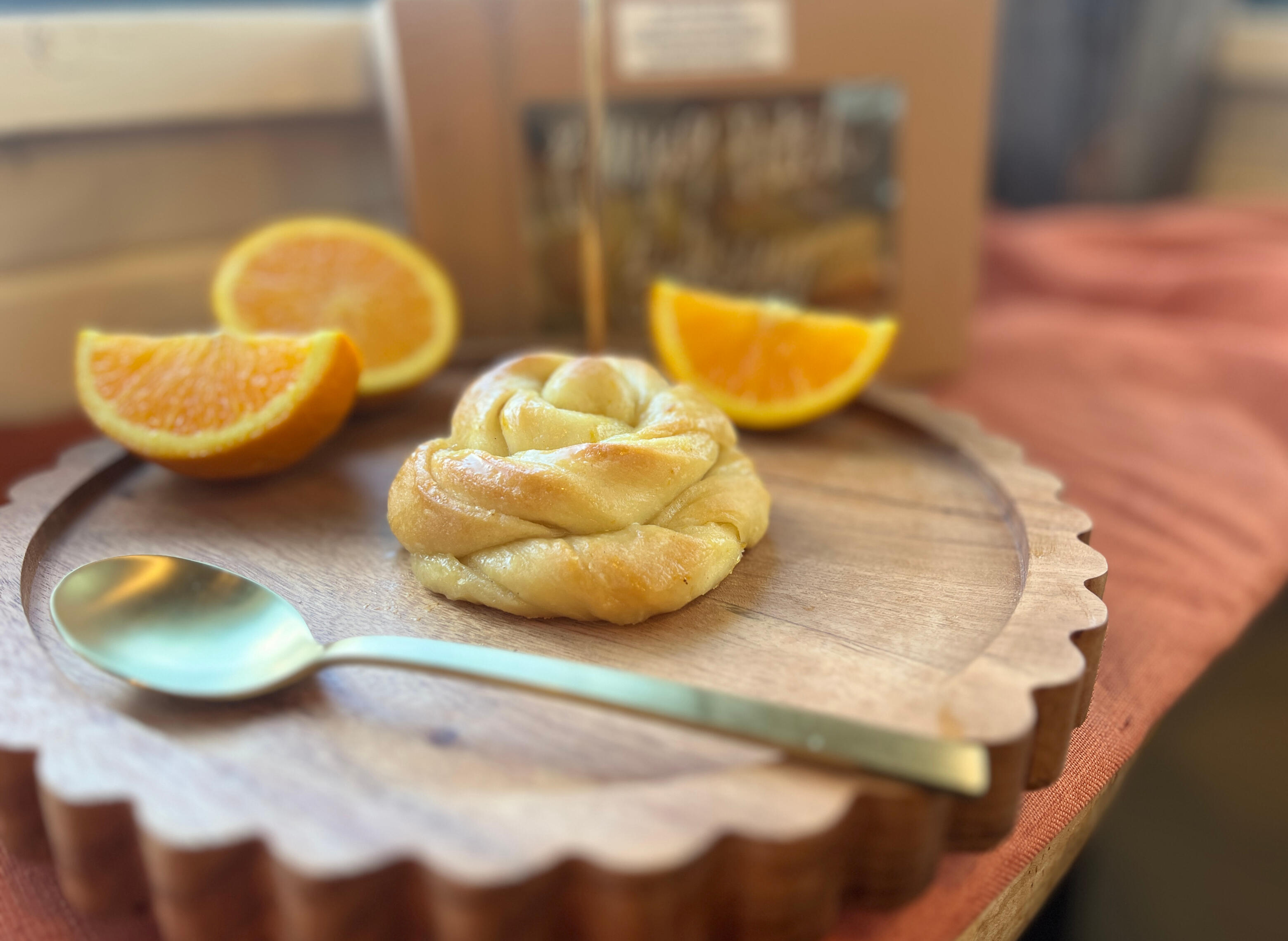 Image: A blossom bun, a pale gold bread roll twisted into a rosette shape and glistening with glaze. It sits on a round wooden display with orange slices and a golden spoon.