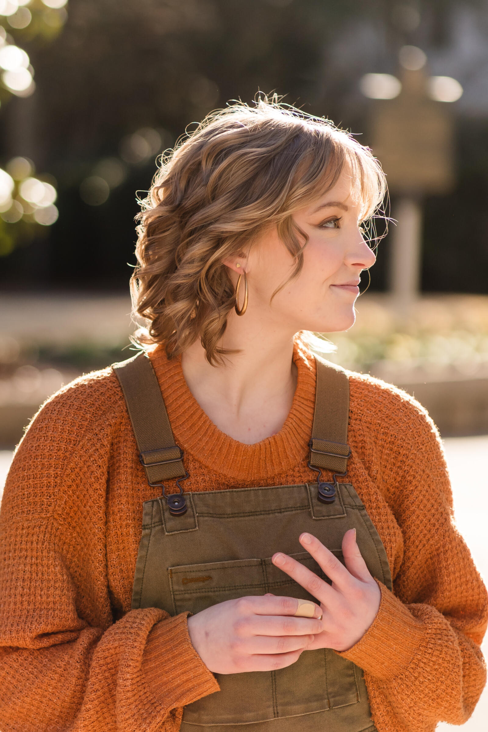 Image: The baker, a young white AFAB with short, curled blonde hair and bangs. They are wearing an orange sweater, green overalls, and gold earrings, and looking off to the left.